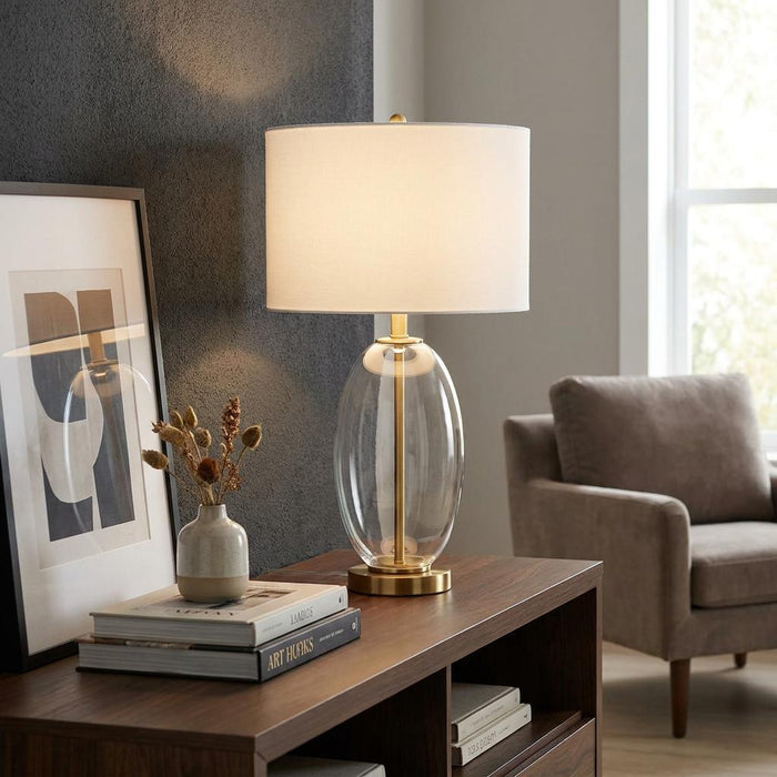 Warmly lit brass table lamp with an oval glass base and white drum shade on a wooden console.