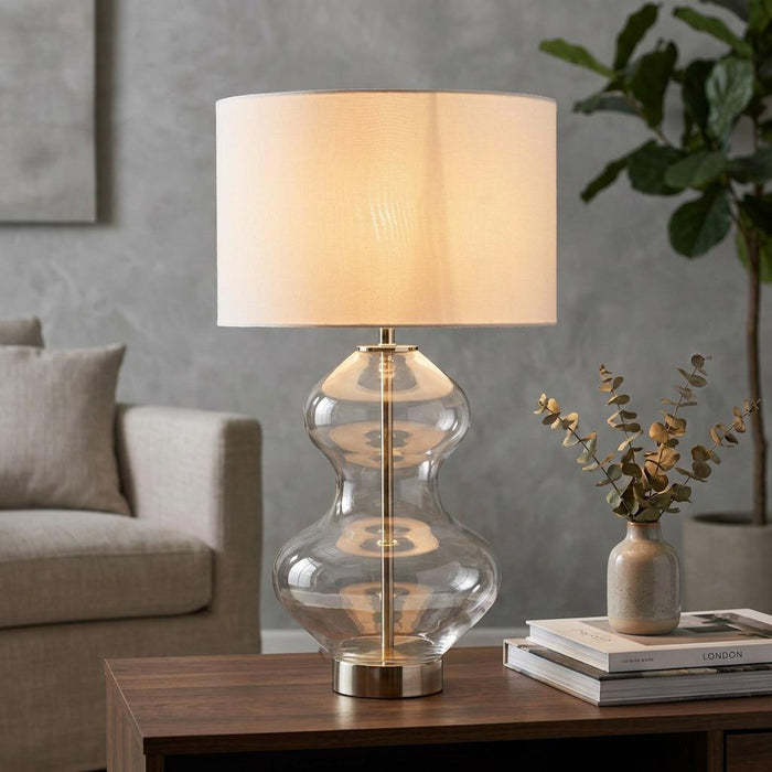 Large touch table lamp with a curved clear glass base and white drum shade on a wooden table in a cozy living room.