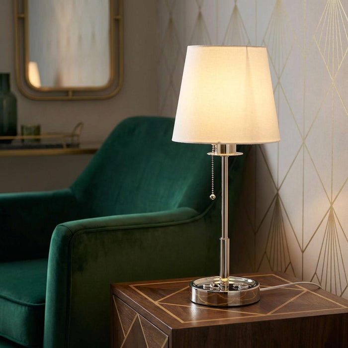 Nickel Art Deco table lamp with a white shade on a wooden table beside a green velvet armchair.