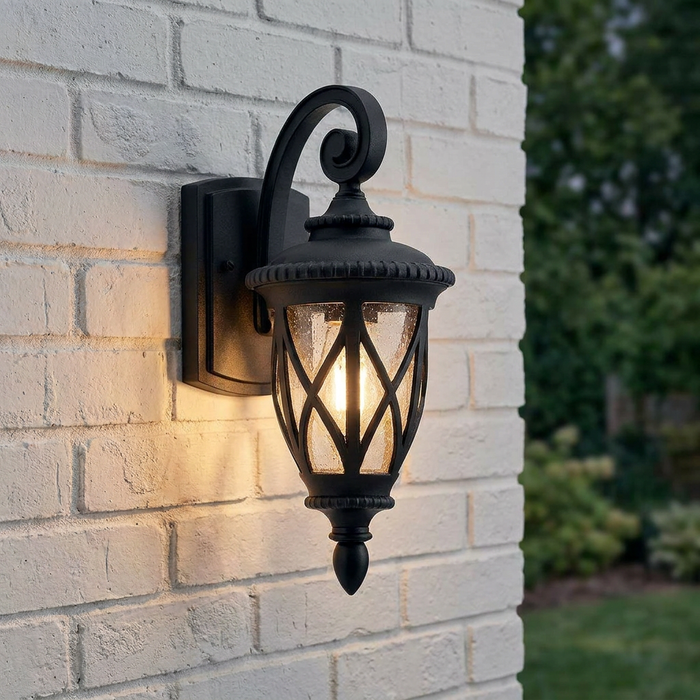 Black outdoor lantern wall light with an illuminated bulb and scroll arm mounted on a white brick wall.