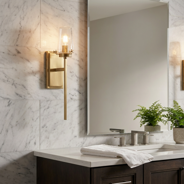 Brushed brass wall light with a clear seeded glass shade mounted on a white marble wall in a bathroom.