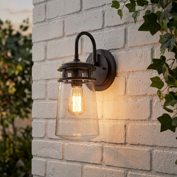 Bronze outdoor wall light with a seeded glass shade and glowing Edison bulb mounted on a white brick wall.
