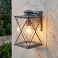 Weathered zinc outdoor wall lantern with X-frame design and seeded glass, glowing on a light stucco wall.