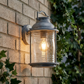 Lit weathered zinc outdoor wall lantern with seeded glass mounted on a brick wall surrounded by green ivy.