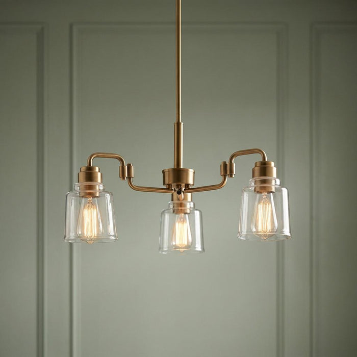 Kichler Avian three-light brass chandelier with clear glass shades and Edison bulbs against a green paneled wall.