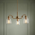 Kichler Avian three-light brass chandelier with clear glass shades and Edison bulbs against a green paneled wall.