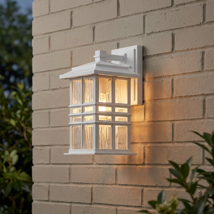 White Craftsman-style outdoor wall lantern with hammered glass panes glowing against a beige brick wall.