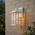 White Craftsman-style outdoor wall lantern with hammered glass panes glowing against a beige brick wall.