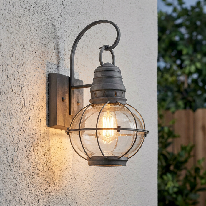 Kichler Bridge Point Zinc Medium Outdoor Wall Light with a caged seeded glass globe mounted on a light stucco wall.