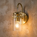 Vintage-style brass wall light with a clear glass jar shade and glowing bulb mounted on a rustic stone wall.
