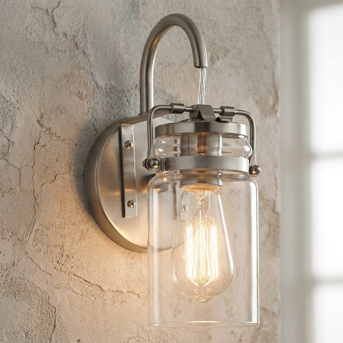 Brushed nickel wall light featuring a clear mason jar glass shade and glowing Edison bulb on a rustic stone wall.