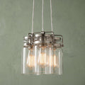 Three-light brushed nickel pendant with glass jar shades and glowing Edison bulbs against a green wall.