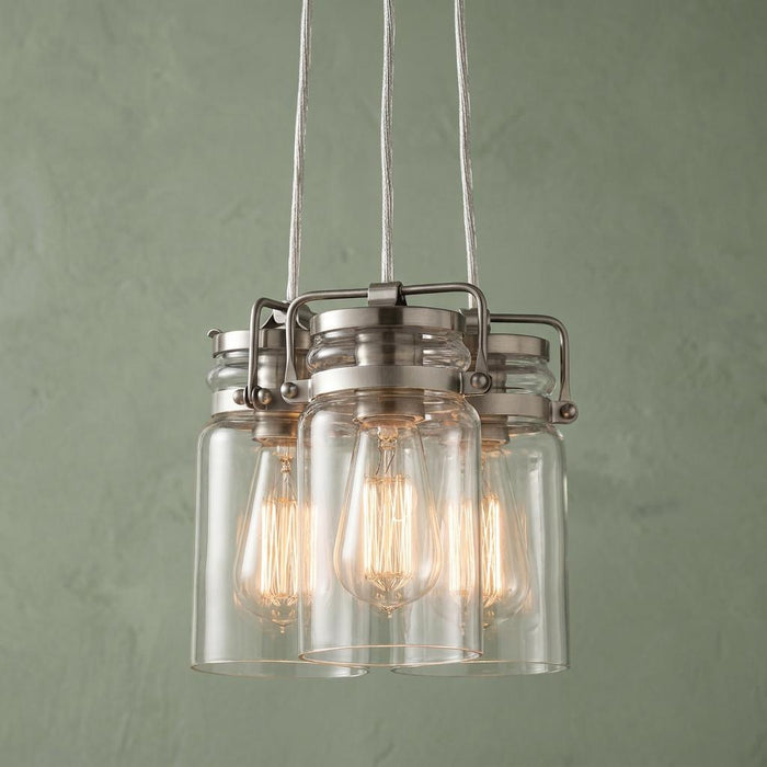 Three-light brushed nickel pendant with glass jar shades and glowing Edison bulbs against a green wall.