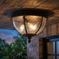 Dark bronze outdoor flush mount ceiling light with seeded glass and glowing bulbs on a wooden porch ceiling.