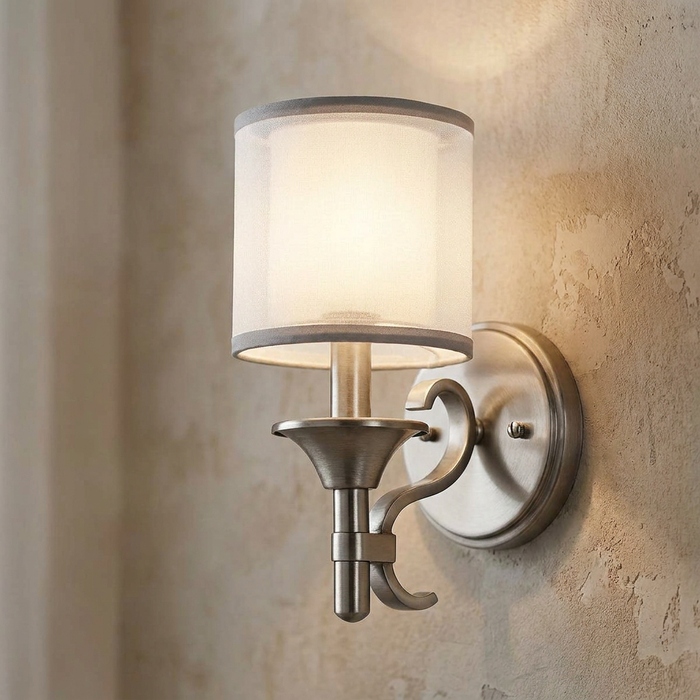 Antique pewter wall light with a white fabric shade mounted on a textured beige wall, emitting a warm glow.