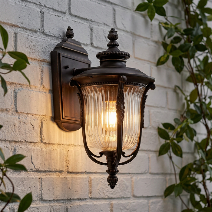 Bronze outdoor wall lantern with clear ribbed glass glowing on a white brick wall with green vines.