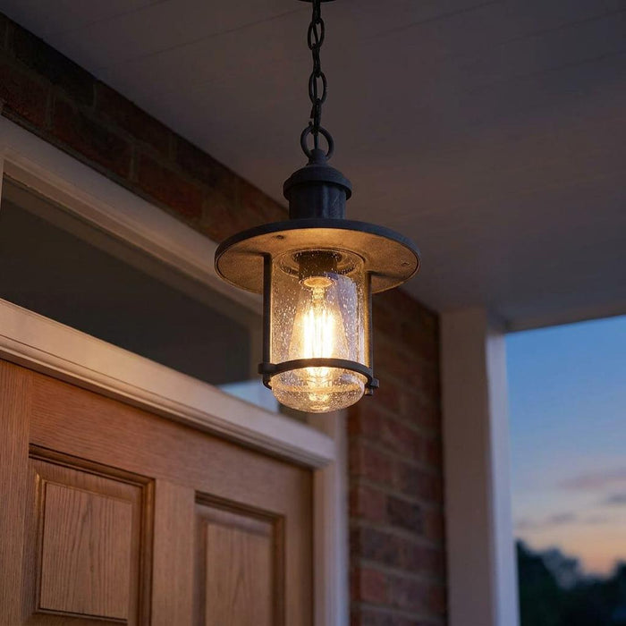 Zinc outdoor pendant lantern with seeded glass shade glowing warmly above a wooden door at dusk.