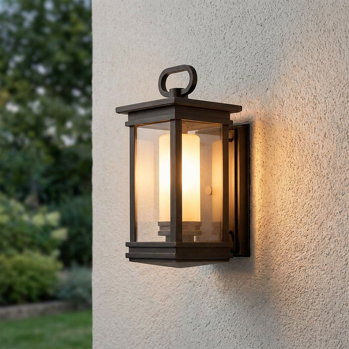 Bronze lantern outdoor wall light with an illuminated frosted glass cylinder on a textured white wall.
