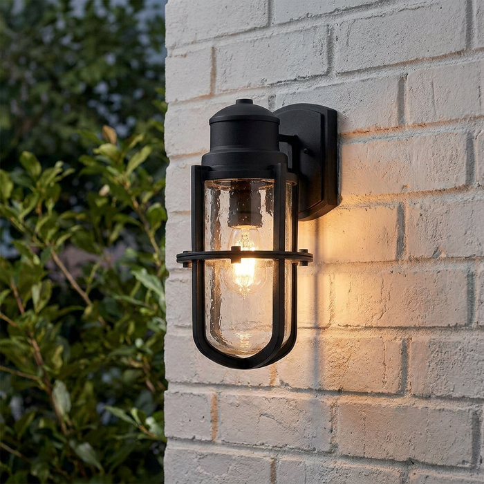 Kichler black outdoor wall light with a glass cylinder and cage frame, mounted on a white brick wall and lit.