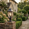 An illuminated black outdoor pedestal lantern sits on a brick pillar in a lush garden pathway.