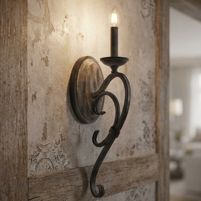 Weathered zinc one-light wall sconce with a scroll design and candle-style bulb on a rustic textured wall.