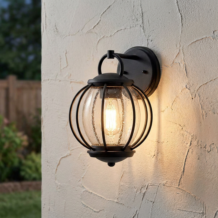 Black outdoor wall light with a seeded glass globe glowing on a textured stucco wall at dusk.