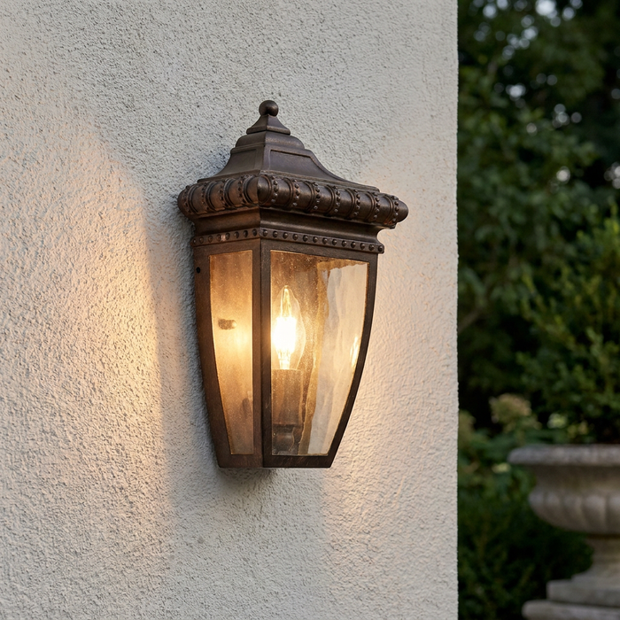 Ornate brushed bronze outdoor wall lantern with clear seeded glass panels and a glowing bulb on a textured wall.