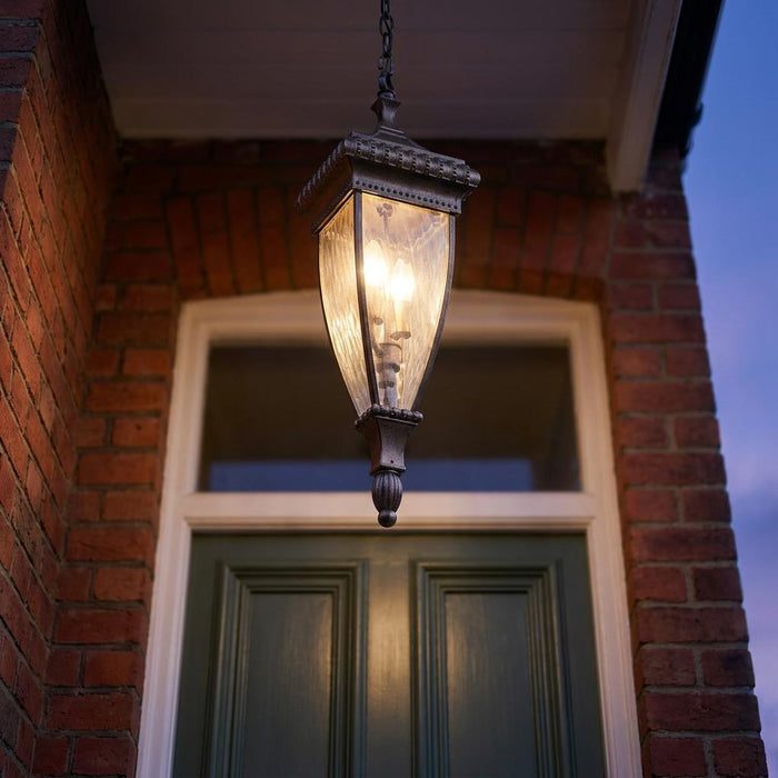 Illuminated brushed bronze outdoor pendant lantern hanging in front of a green door under a brick porch.
