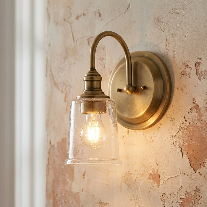 Kichler Waverly brass wall light with a seeded glass shade and warm bulb on a distressed plaster wall.