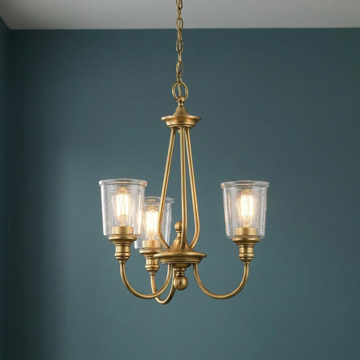 Three-light natural brass chandelier with clear seeded glass shades against a teal wall.