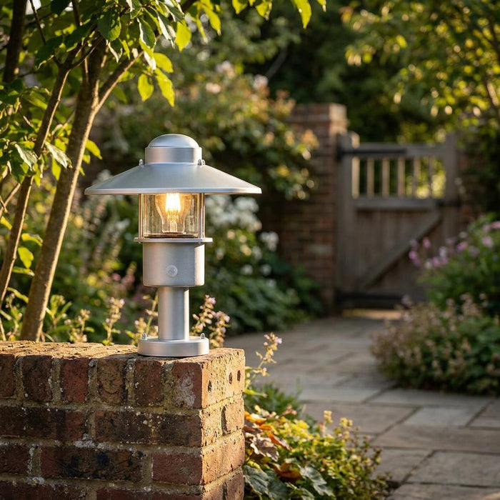 Silver industrial-style pedestal light glowing on a brick wall in a lush garden setting.