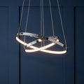 Modern chrome pendant ceiling light with two interlocking LED rings against a dark blue paneled wall.