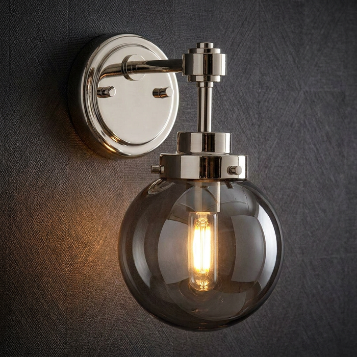 Polished nickel wall light with a smoked glass globe and glowing bulb mounted on a dark textured wall.