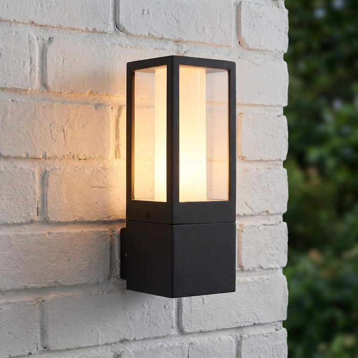 Modern black rectangular outdoor wall lantern with a glowing cylindrical diffuser mounted on a white brick wall.