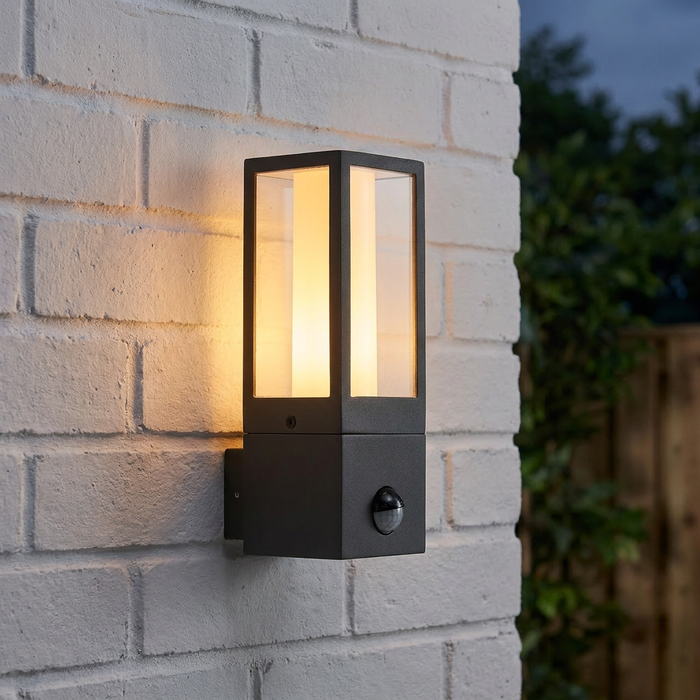 Grey rectangular outdoor lantern with a warm internal glow and PIR sensor, mounted on a white brick wall at dusk.
