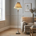 Traditional floor lamp with a soft grey pleated shade glowing warmly next to an armchair in a neutral living room.