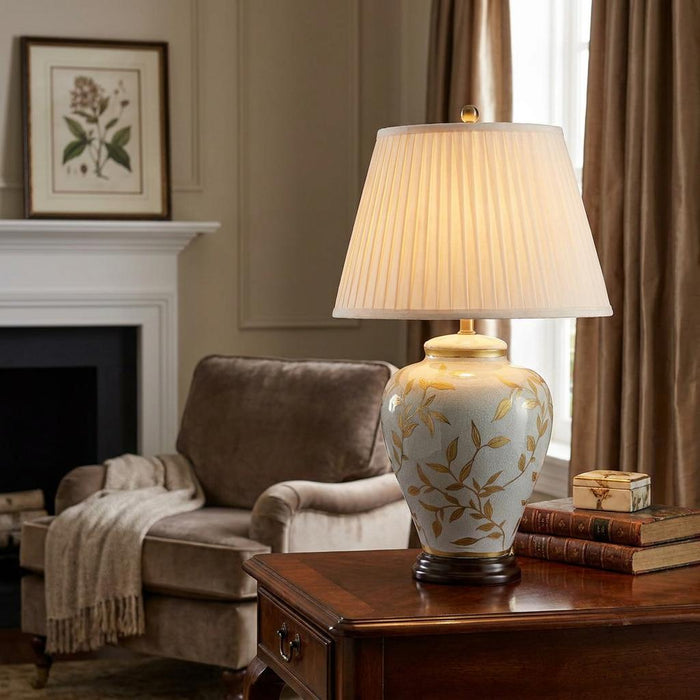 Ceramic table lamp with gold leaf patterns and a pleated shade on a wooden table in a cozy living room.