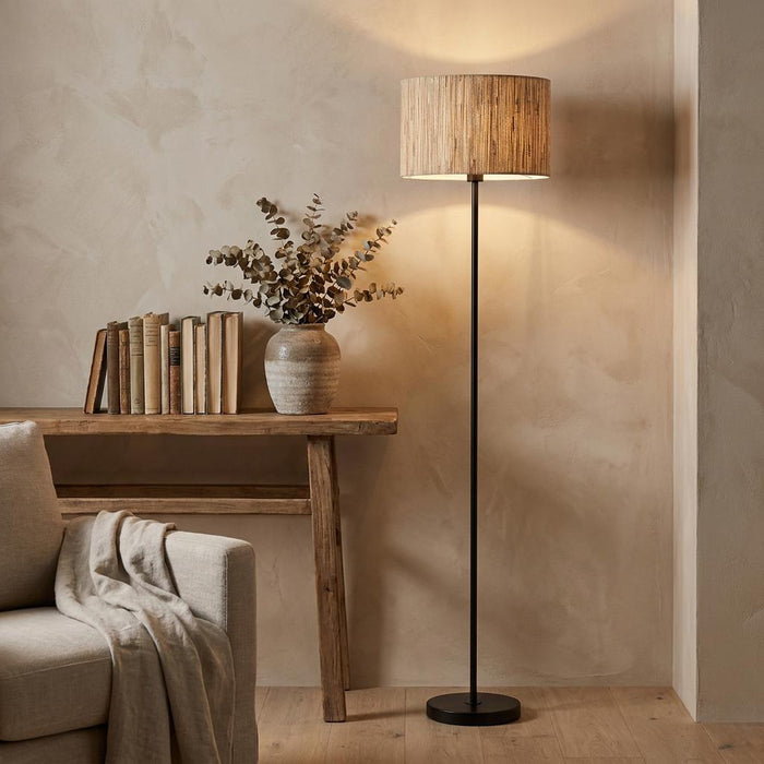 Black floor lamp with a woven drum shade illuminated in a neutral-toned room next to a rustic wooden table.
