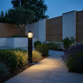 Black louvre bollard light glowing warmly beside a stone garden path at twilight.