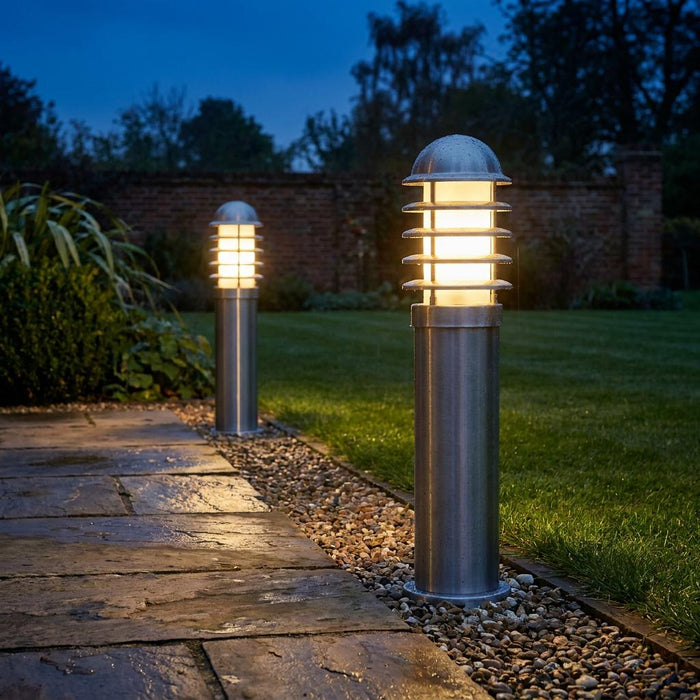 Two illuminated stainless steel louvre bollard lights lining a wet stone garden path at twilight.