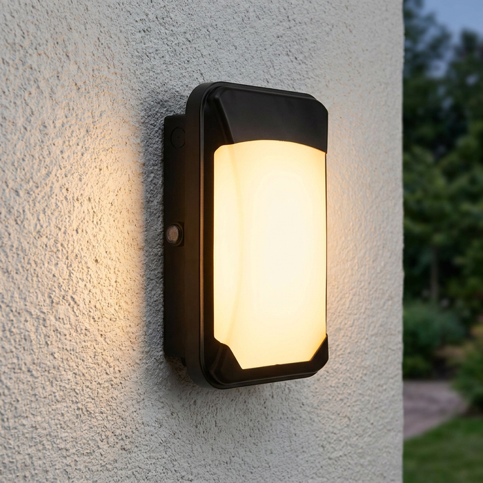 Rectangular black LED outdoor wall light with a frosted diffuser glowing warmly on a white textured wall.