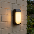 Rectangular black outdoor wall light with a frosted lens emitting a warm glow, mounted on a brick wall.