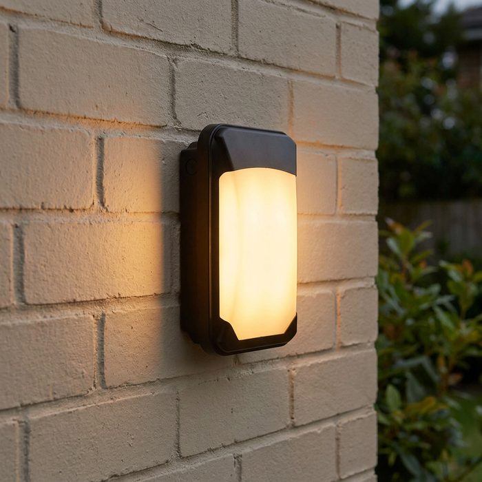 Rectangular black outdoor wall light with a frosted lens emitting a warm glow, mounted on a brick wall.