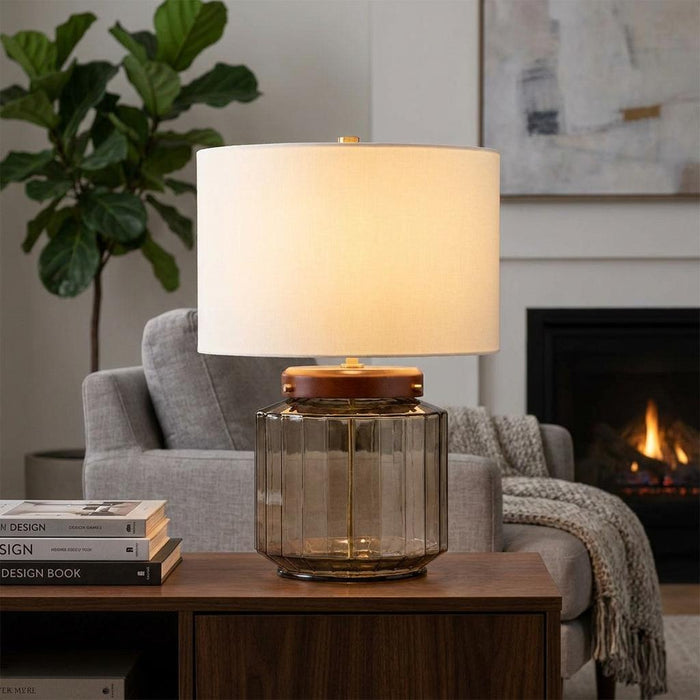 Lit Luga table lamp with ribbed smoke glass base and white drum shade in a cozy living room setting.
