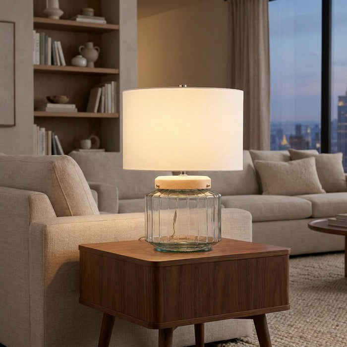 Illuminated Luga glass table lamp with a white drum shade on a wooden side table in a modern living room.