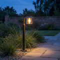 Lutec Cate outdoor bollard light in black, glowing warmly at twilight beside a stone pathway and garden shrubbery.