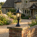 Traditional black lantern outdoor light illuminated on a brick garden wall pillar in front of a stone cottage.