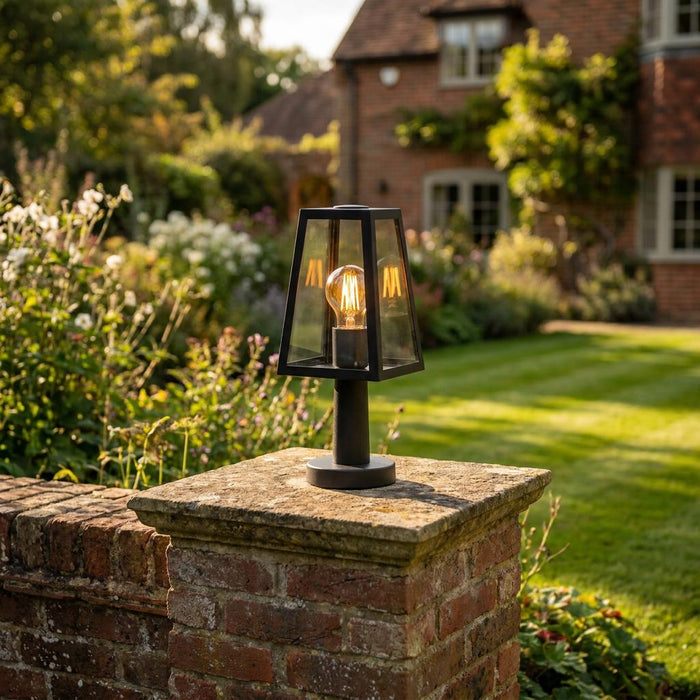 Black lantern-style garden light with a warm Edison bulb mounted on a brick post in a sunny garden.