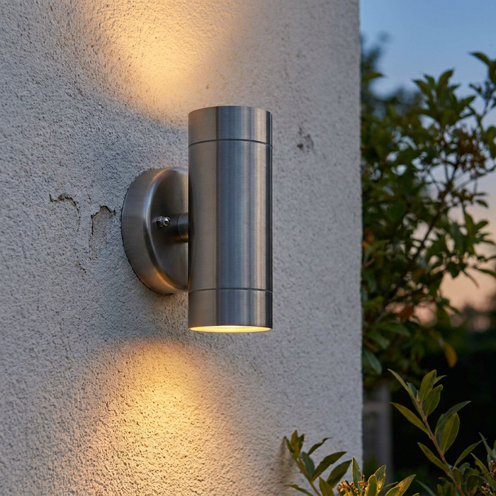 Cylindrical stainless steel up and down wall light illuminating a textured exterior wall with warm light at dusk.