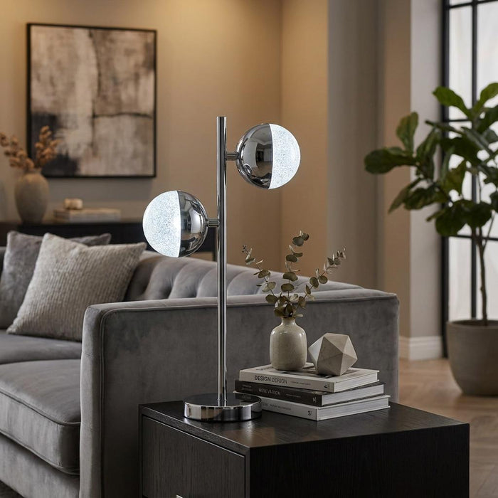 Chrome table lamp with two crystal sand glass globes on a side table in a modern living room setting.